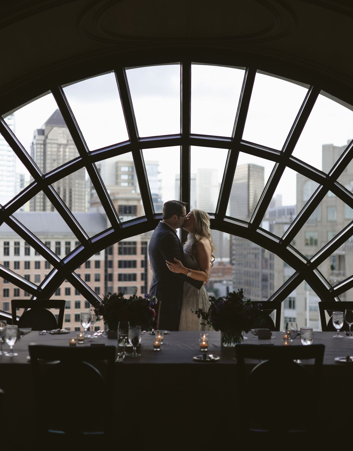 Couple kissing in silhouette before arched window with city skyline backdrop at elegant urban wedding reception.