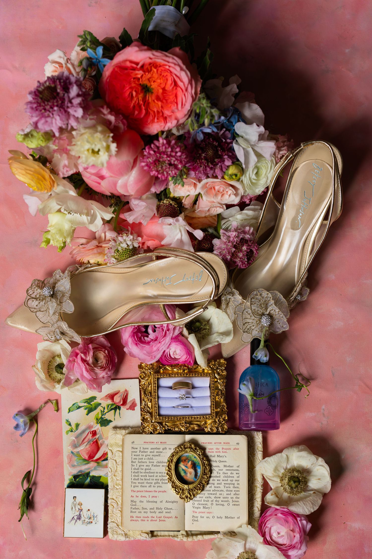 Bridal flat lay with gold heels, floral bouquet, wedding rings, vintage book, and decorative accessories on pink background.