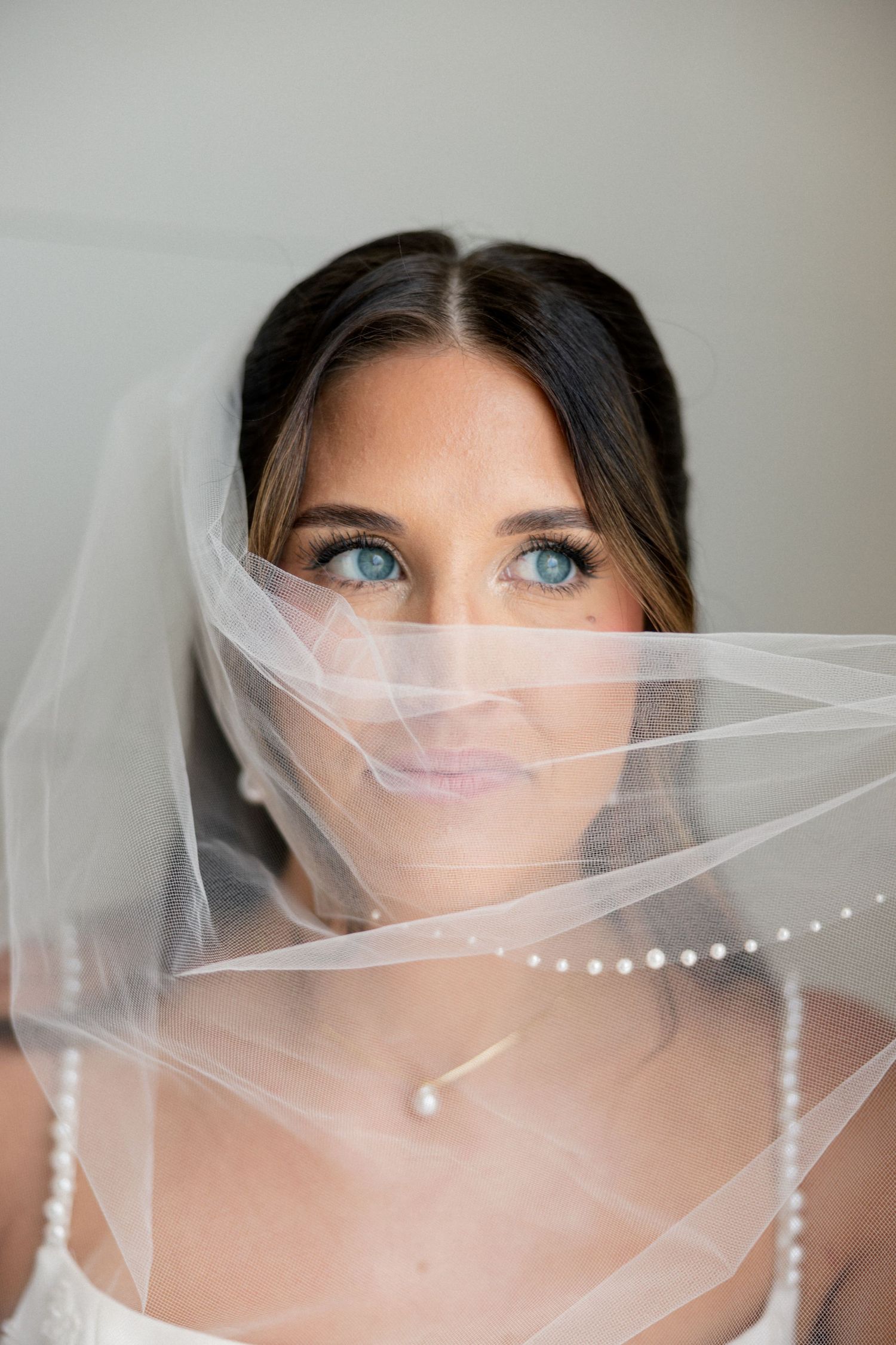 A bride with blue eyes gazes upward through a flowing pearl-trimmed white veil, wearing a delicate necklace.