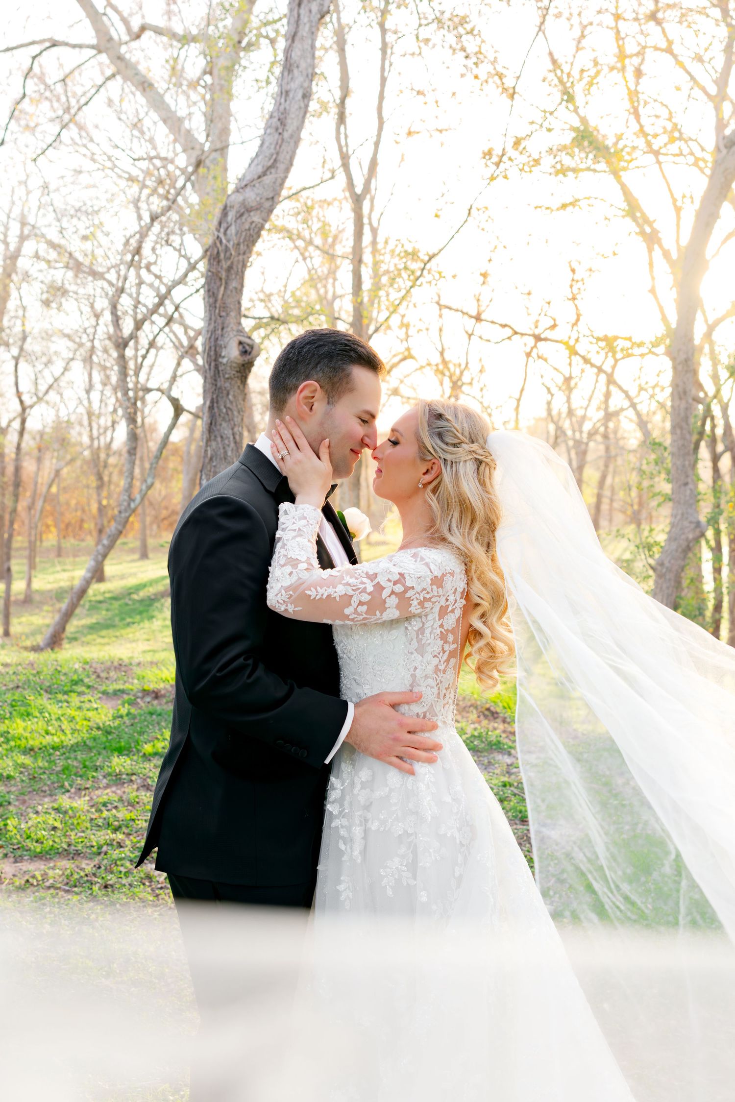 Bride and groom share a romantic almost-kiss outdoors, her lace veil flowing, surrounded by golden sunlit trees.