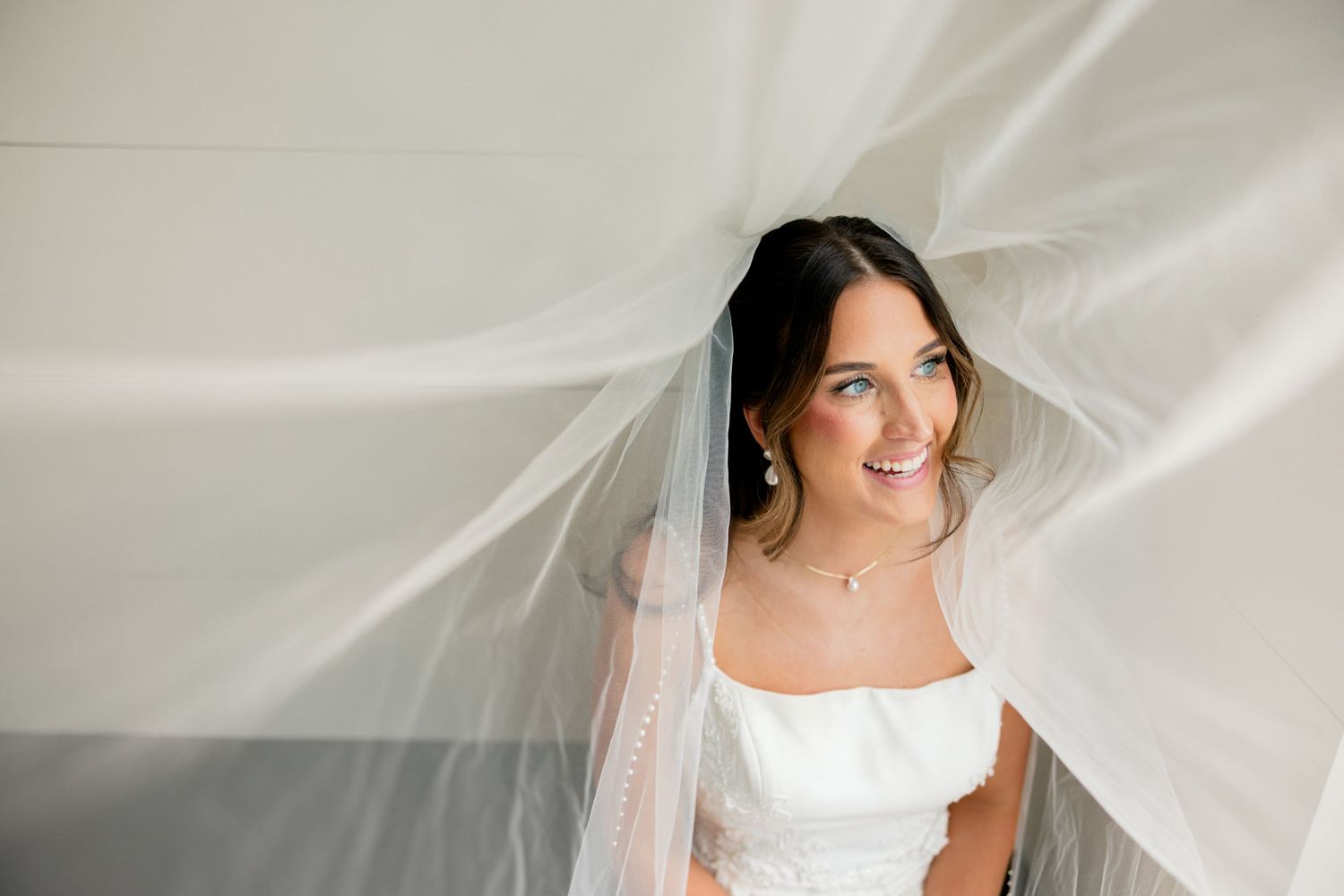 Bride in white gown smiling under flowing veil, wearing elegant jewelry, captured in a dreamy bridal portrait.