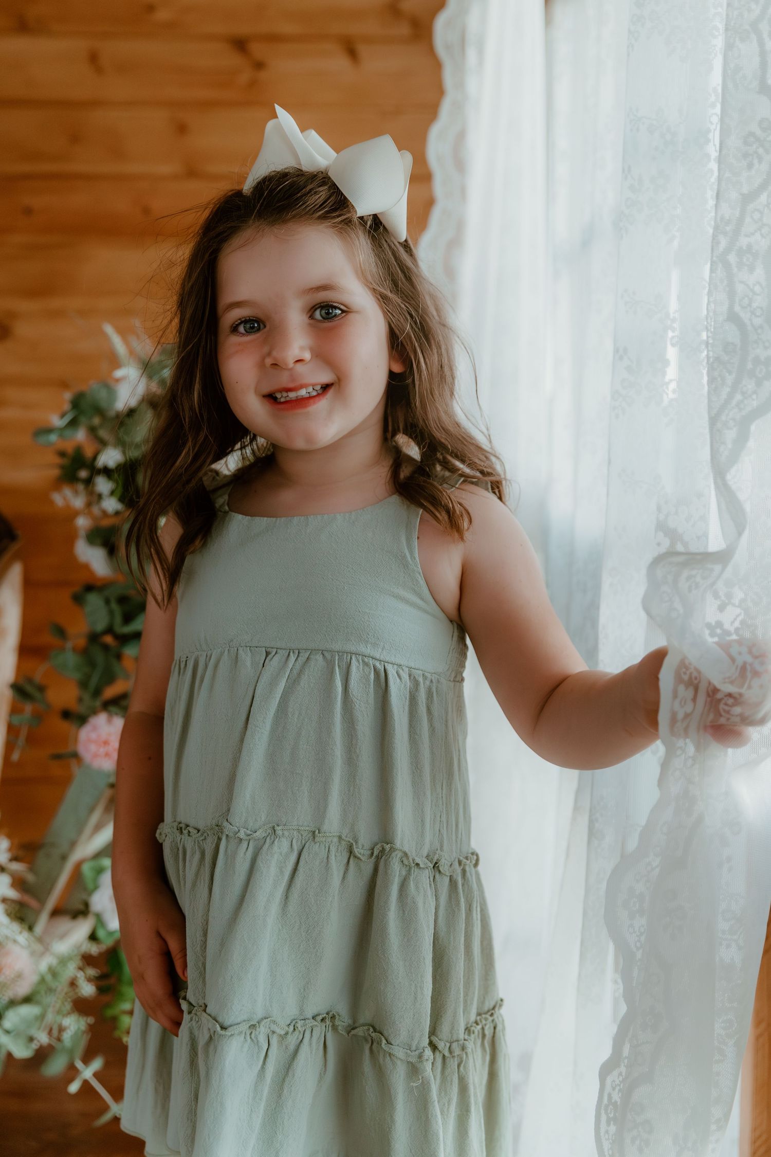 A soft sage green tiered dress with white bow accessory against a rustic wooden wall and lace curtain backdrop.