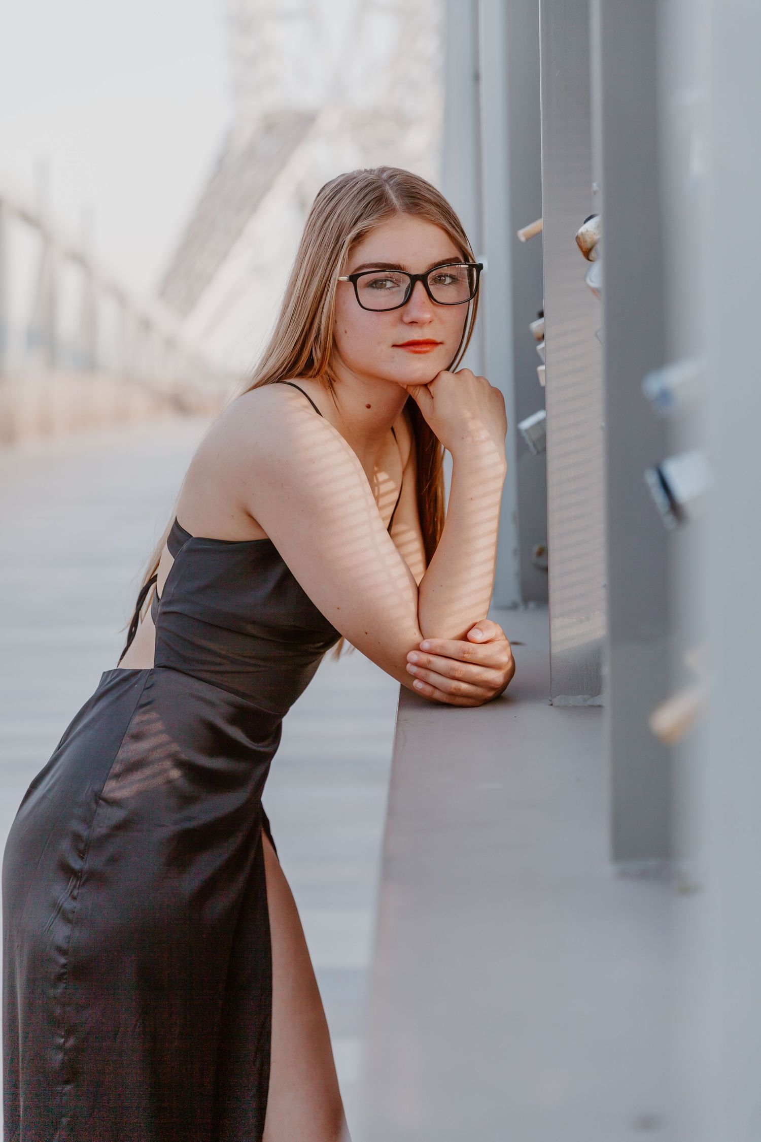 A woman in glasses and a black evening dress leans against a wall in soft lighting.