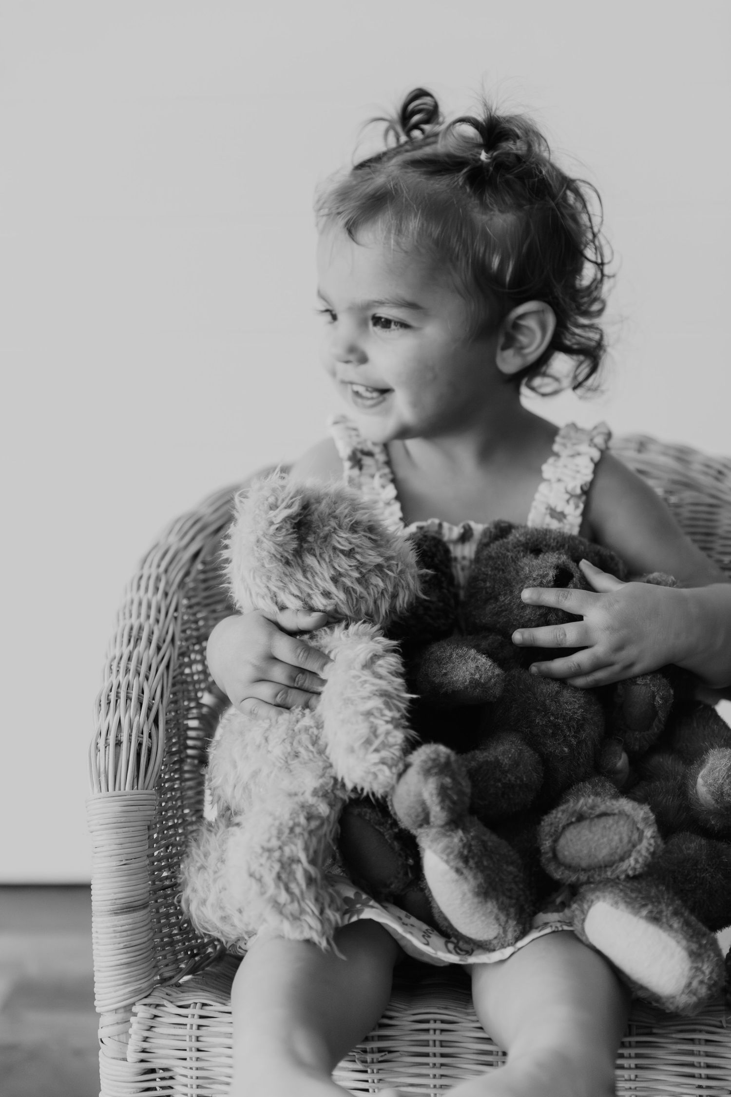 Black and white photo of a small child seated in a wicker chair hugging stuffed teddy bears and smiling.