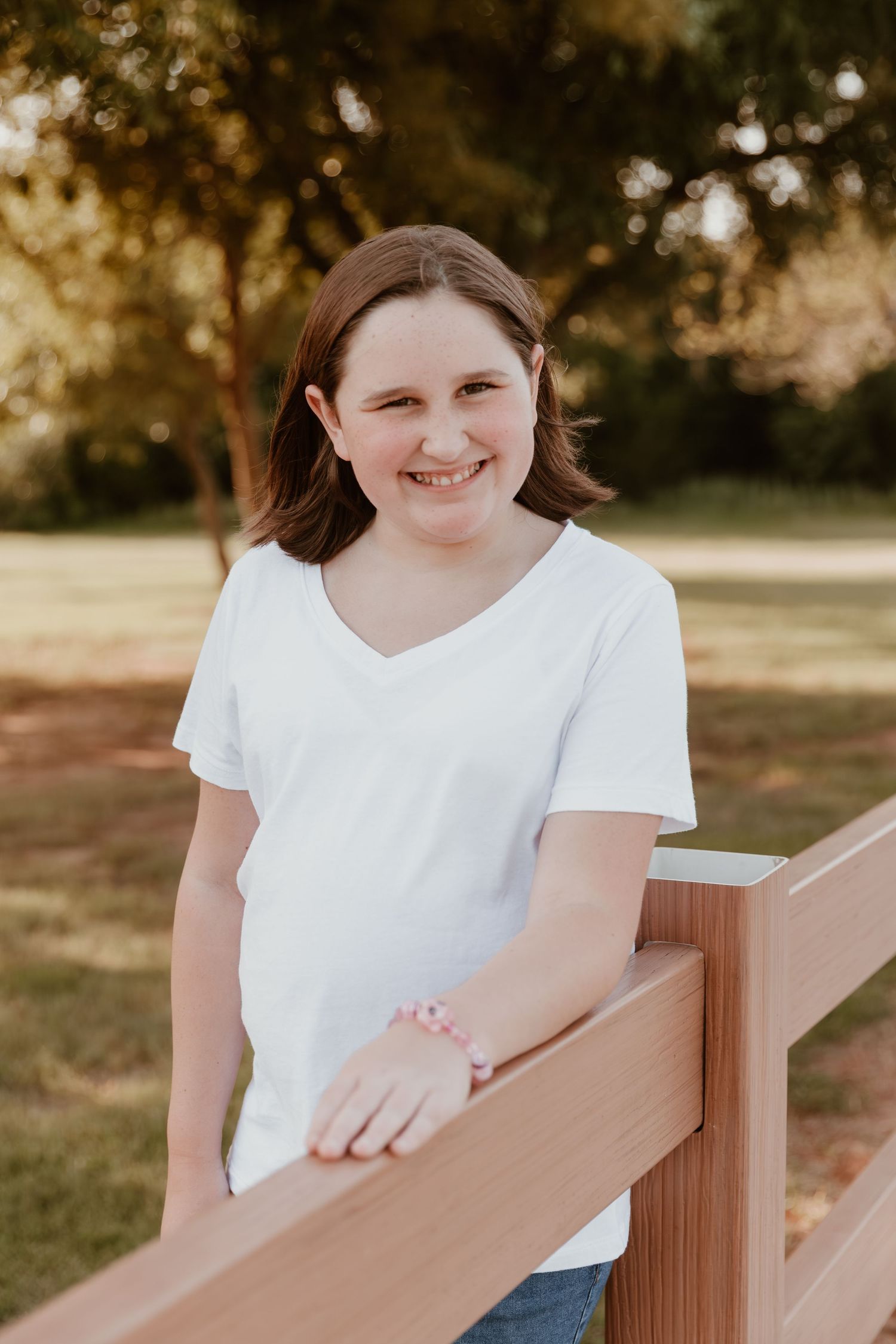 Smiling person in a white v-neck t-shirt leaning on a wooden railing during golden hour in an outdoor setting.