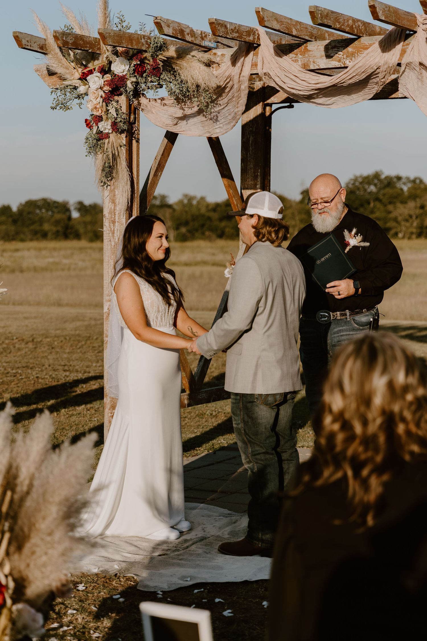 A romantic outdoor wedding ceremony takes place under a rustic wooden arch at sunset in a rural field.