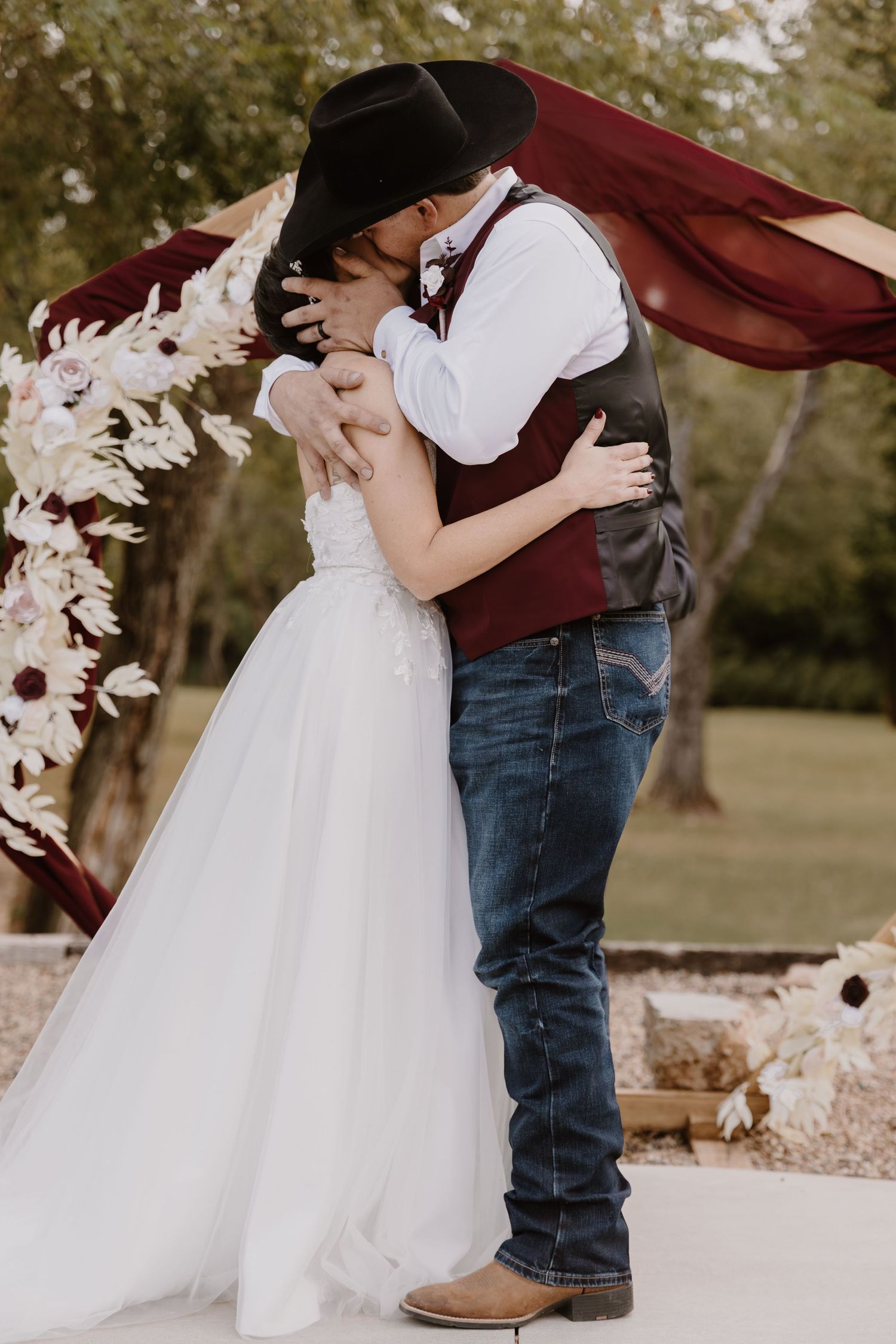A romantic wedding couple shares a kiss under a burgundy arch decorated with white flowers during an outdoor ceremony.