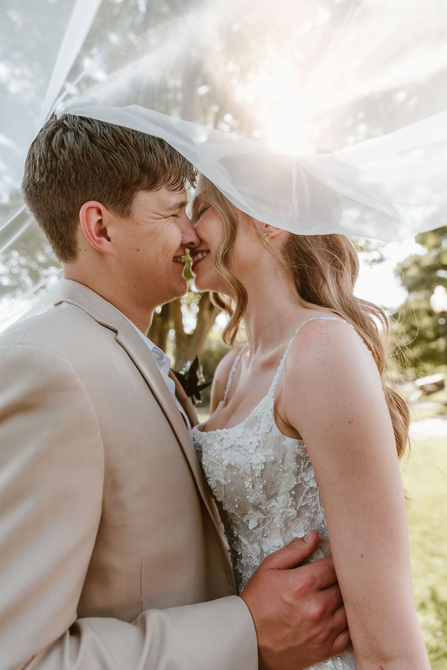 A bride and groom share a romantic moment under a flowing veil, bathed in golden sunlight outdoors.