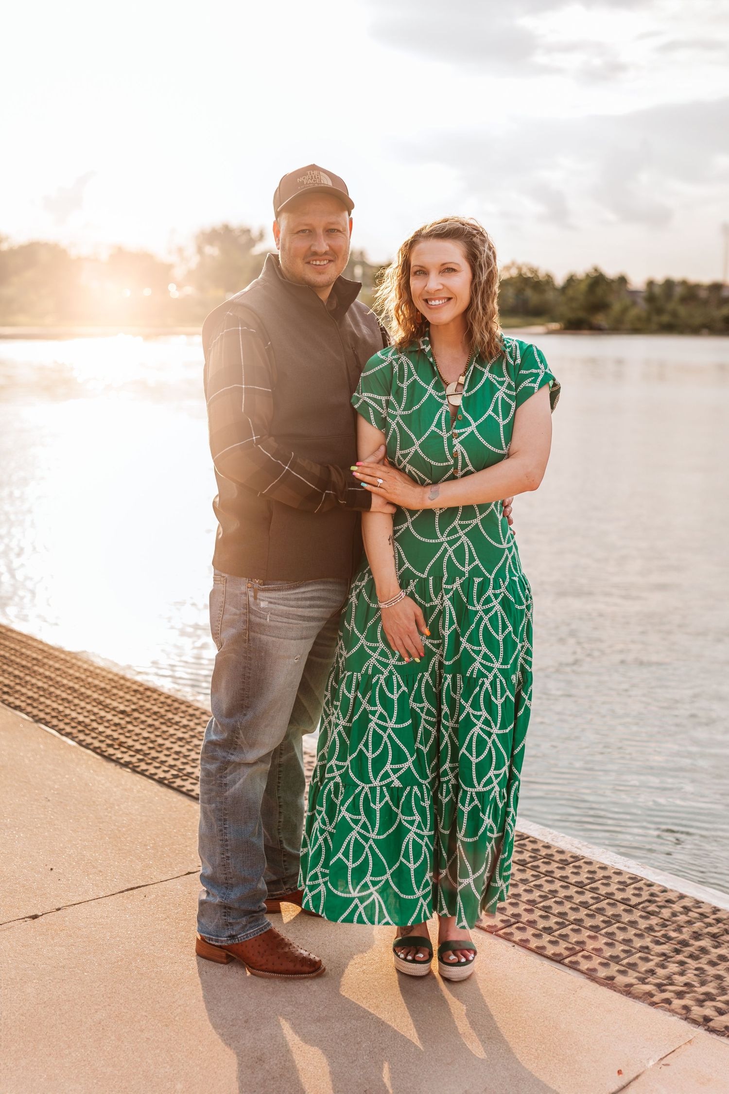 Couple standing by waterfront during sunset, wearing green dress and casual attire.
