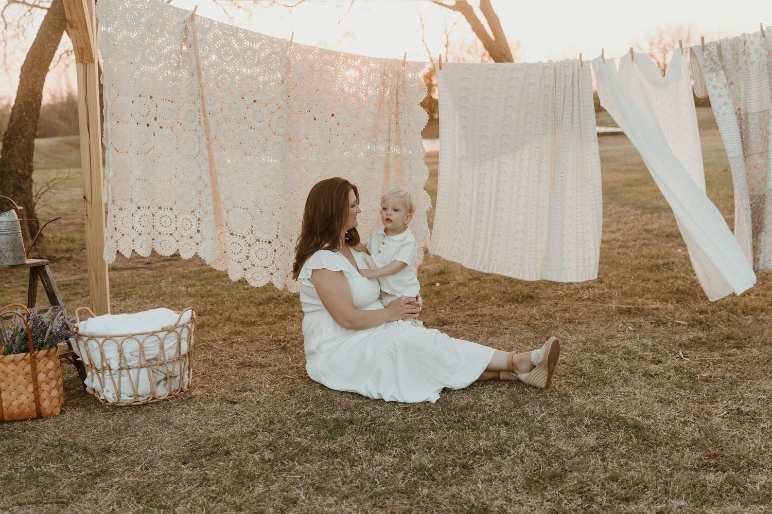 Mother in white dress sitting on grass with baby, surrounded by hanging lace linens outdoors at golden hour.