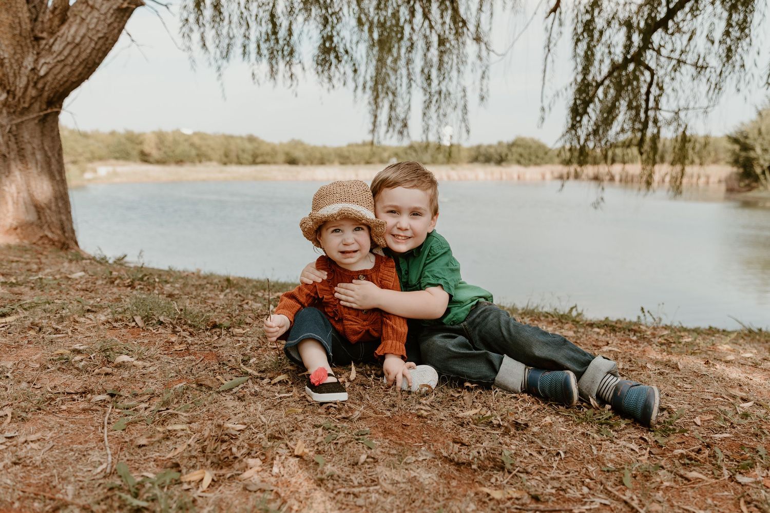 Two children in casual clothes laying on a dirt slope near a lake on a sunny autumn day.