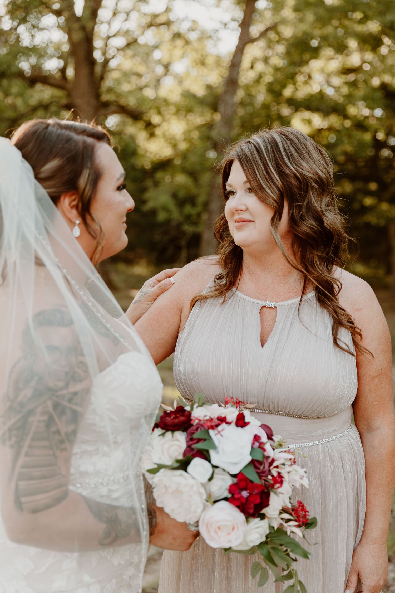 Row of bridesmaids in light gray dresses hold red and pink rose bouquets while sharing candid moments at outdoor wedding.