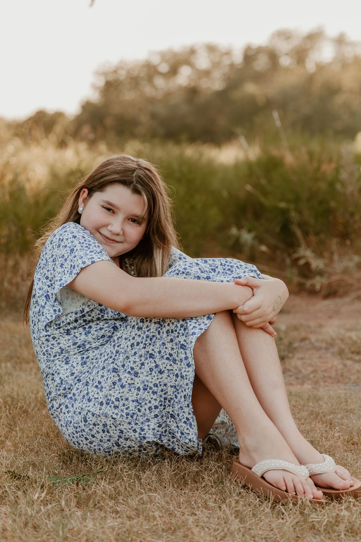 A person sits on dry grass wearing a blue floral dress and sandals during golden hour.