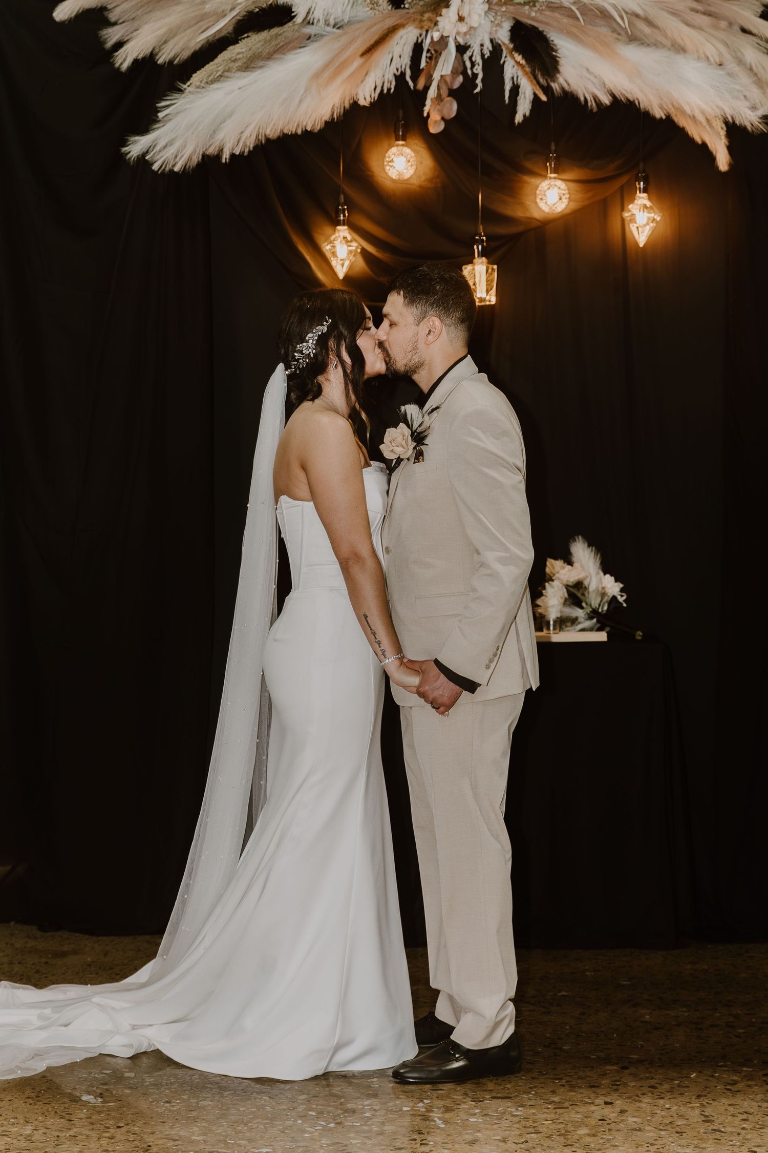 Bride and groom share first kiss at boho wedding altar with pampas grass and Edison bulb string lights.
