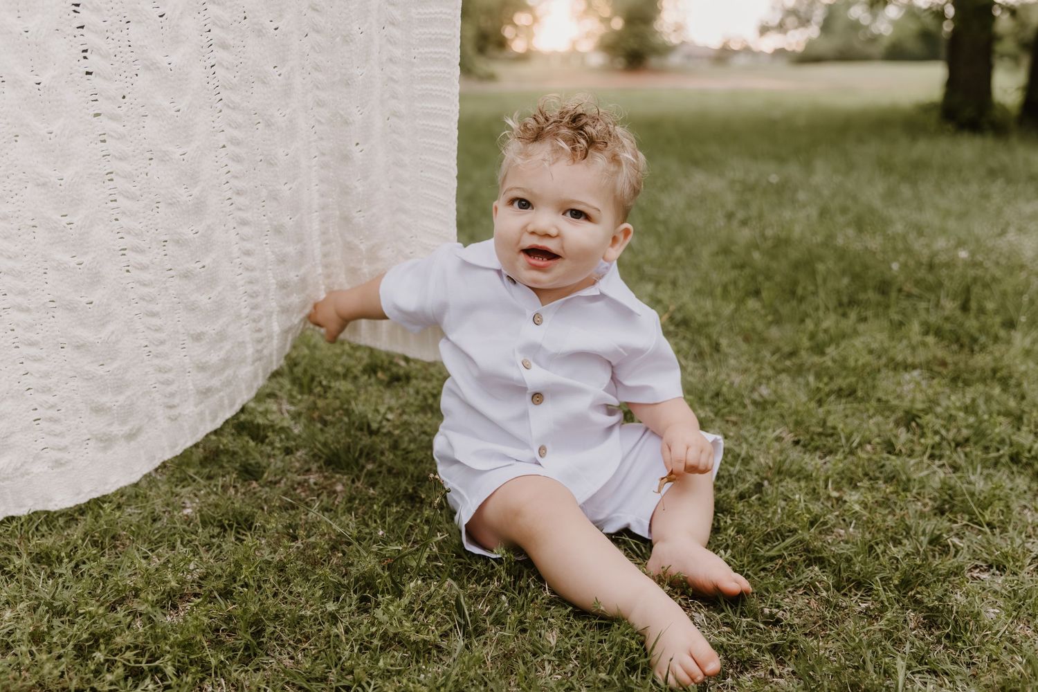 A happy baby boy in a white romper sits on green grass outdoors during a golden hour photo session.