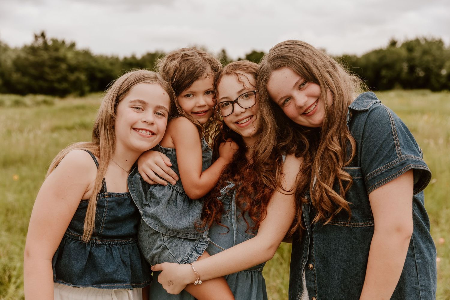 Four girls smiling and hugging outdoors in a green field, wearing denim outfits in a warm family portrait.