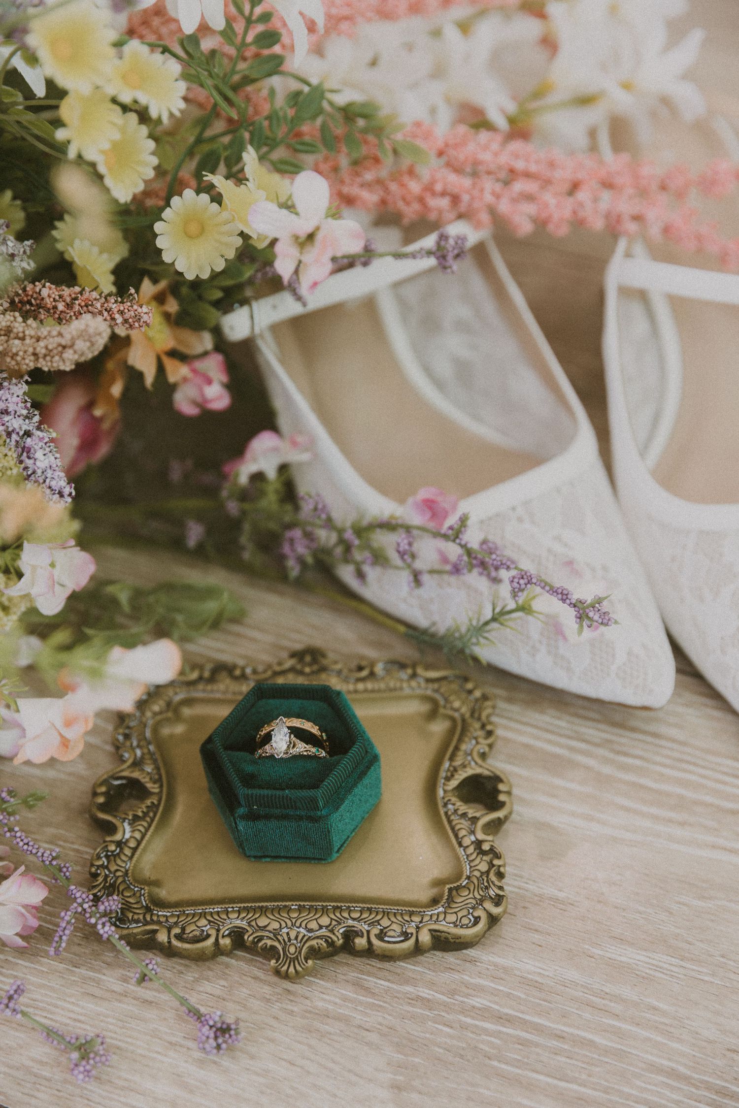 Wedding rings in a green velvet hexagon box on a gold tray, surrounded by wildflowers and lace bridal shoes.