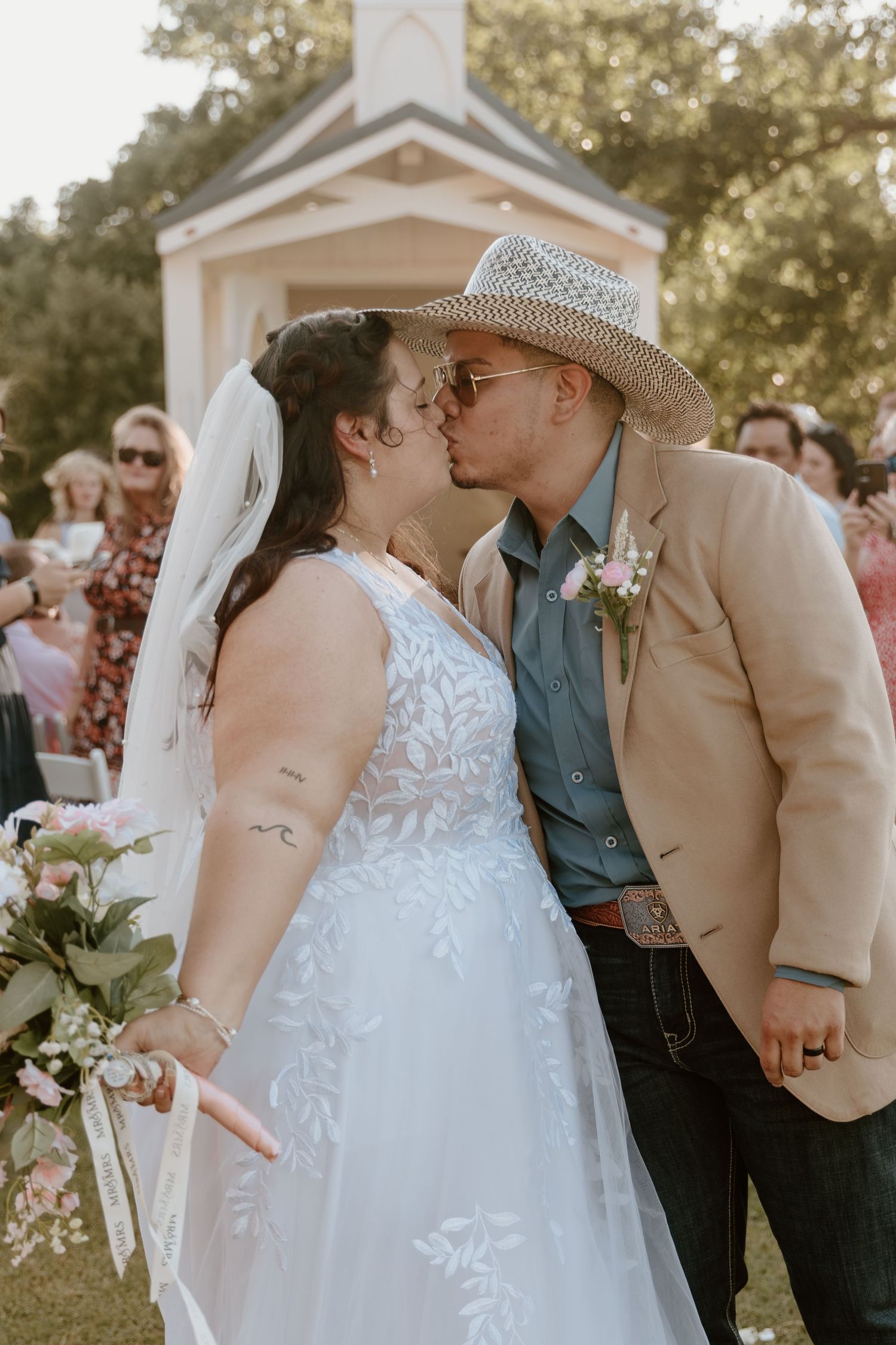 Bride and groom sharing first kiss at outdoor wedding ceremony, groom wearing cowboy hat.