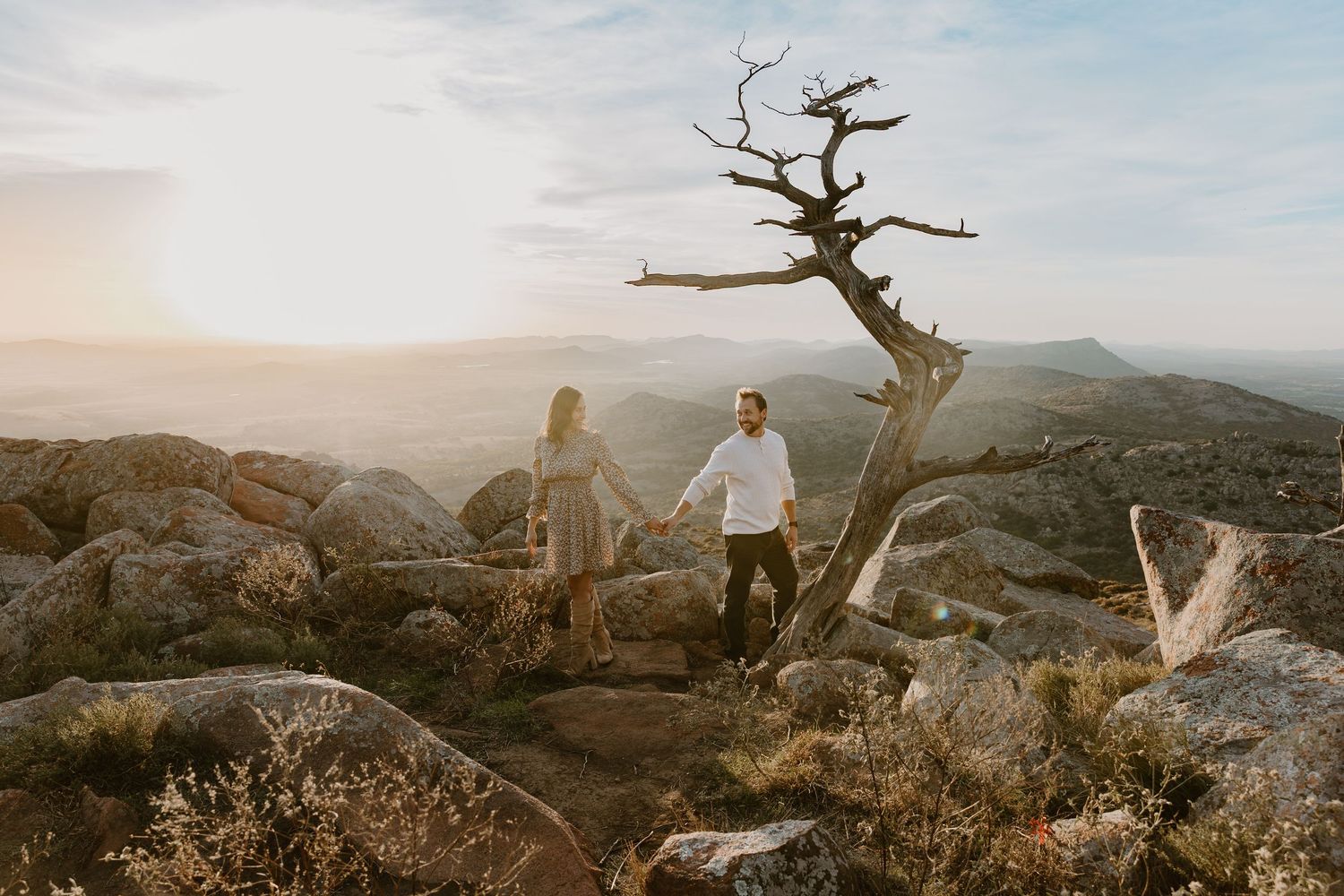 A panoramic mountain landscape view at sunset with silhouetted figures and rocky outcrops against a golden sky.