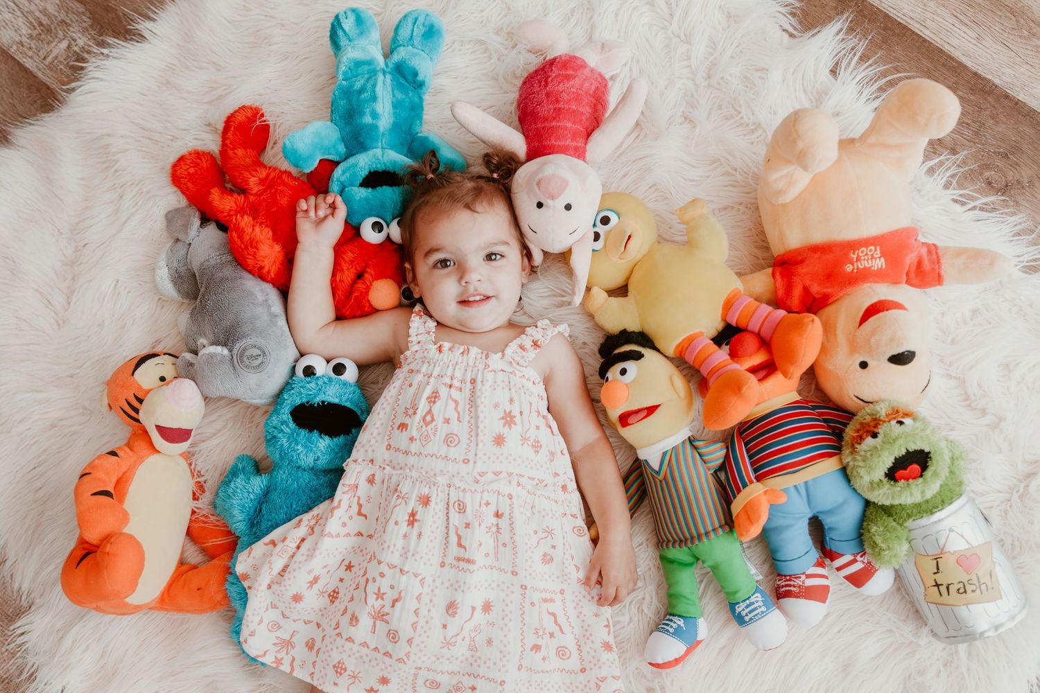 A baby in white dress lies on furry blanket surrounded by colorful stuffed animal toys.