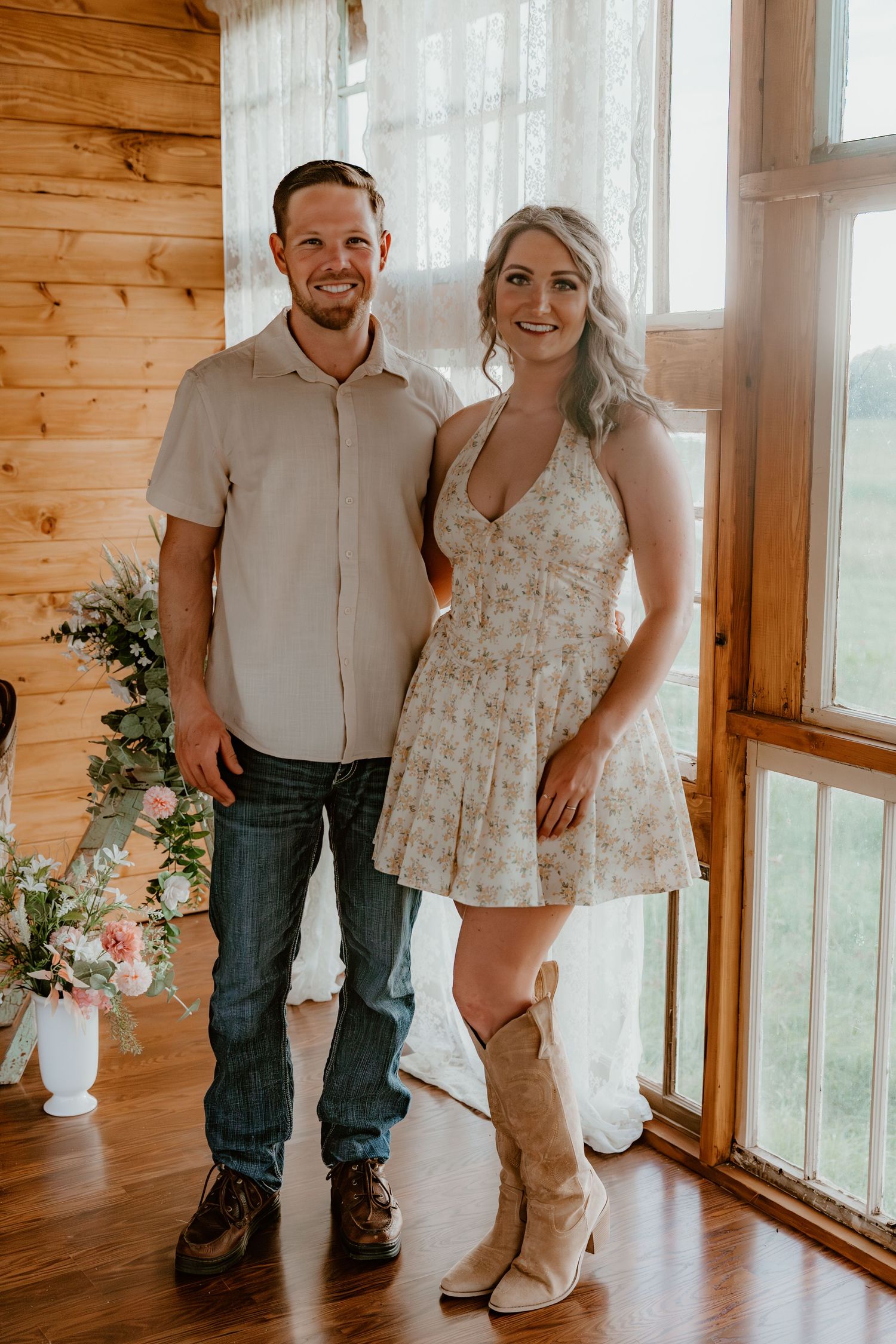 Two people stand together in a rustic wooden room with large windows and warm lighting.