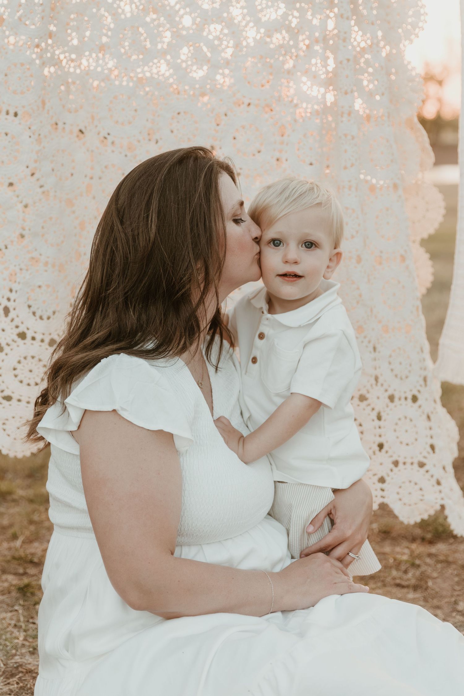 A mother in a white dress kisses her blonde toddler son, both dressed in white, against a lace backdrop outdoors.