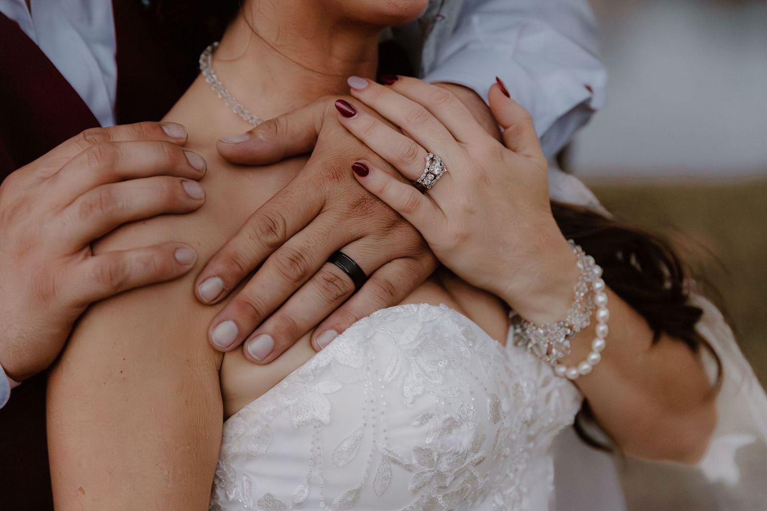 Close-up of bride and groom hands showing wedding rings, pearl bracelet, and dark red nails on white lace dress.