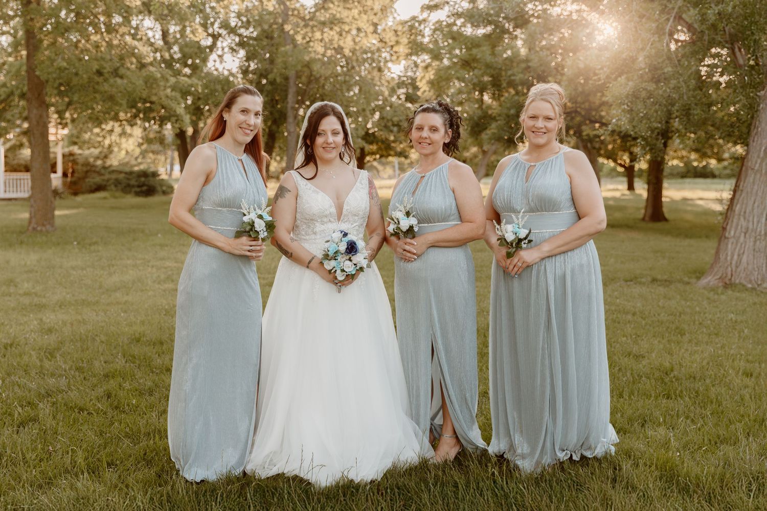 Four people in light blue bridesmaid dresses and white wedding gown stand together in a sunny outdoor setting at sunset.