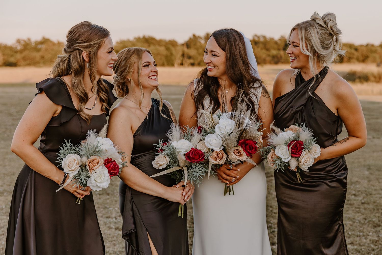Wedding party in brown bridesmaid dresses laughs together while holding white and red bouquets at sunset.