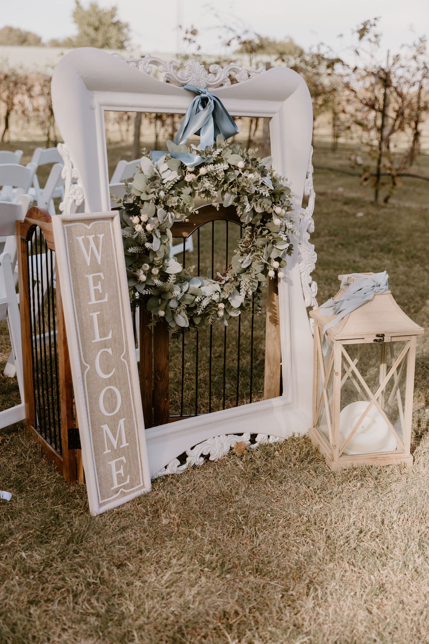 Rustic wedding decor featuring white lanterns with floral accents and 'Welcome' signs arranged on dried grass.