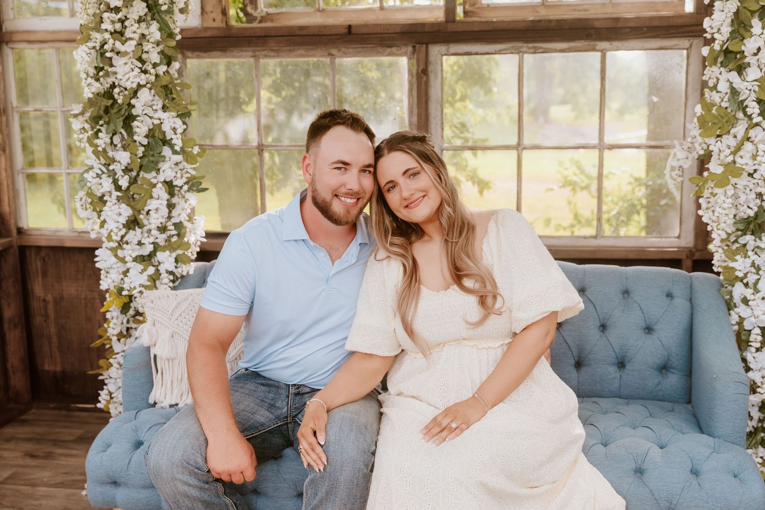 A couple smiles together on a blue tufted sofa surrounded by white flowers in a rustic studio setting.