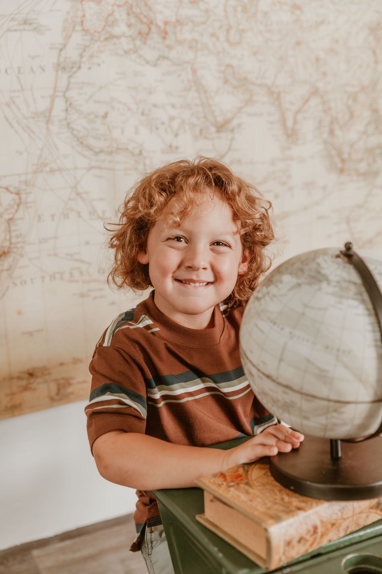 Young child with curly red hair poses next to a vintage globe and books against a map backdrop.