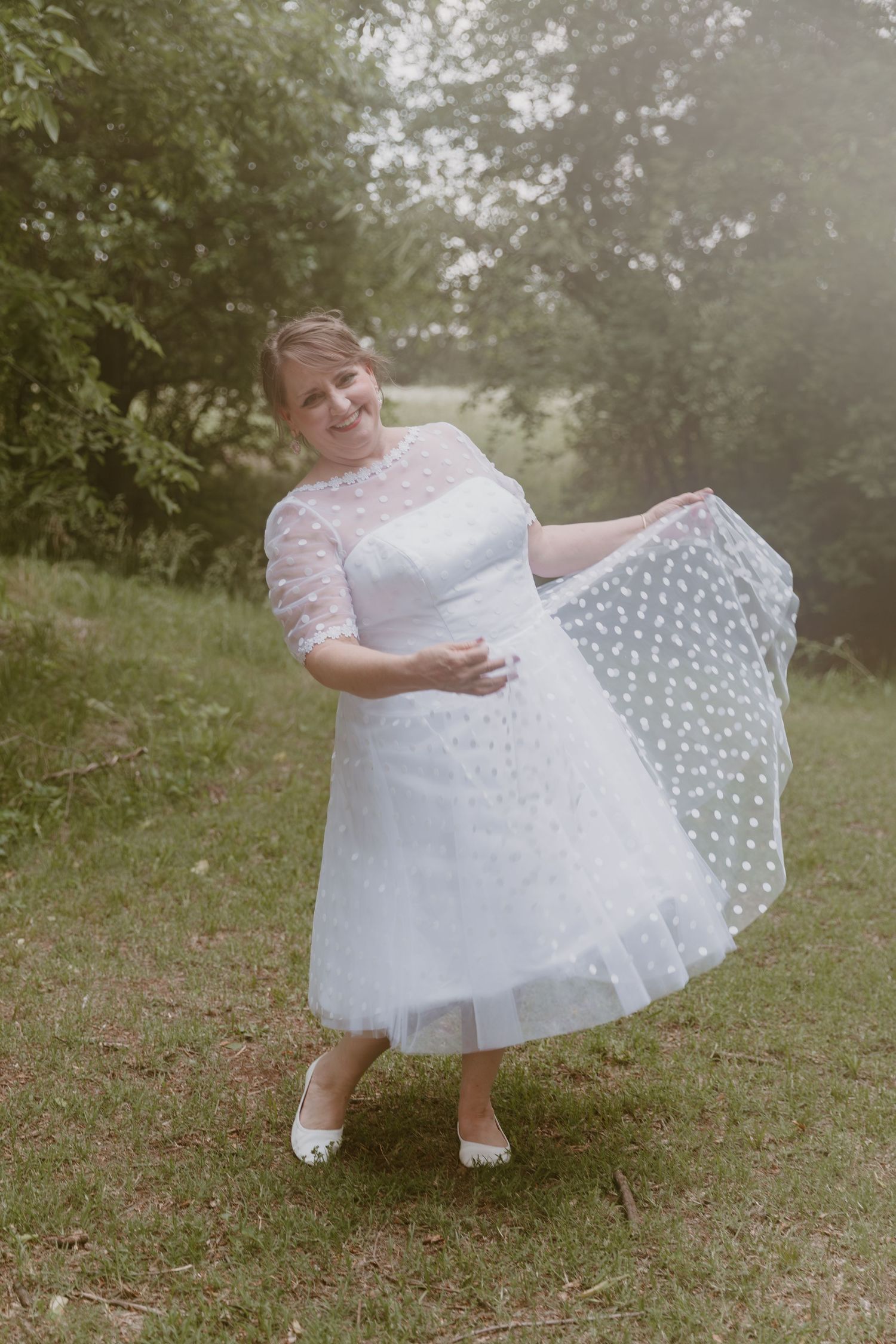 A bride in a tea-length white lace wedding dress with polka dot details twirls outdoors in a garden setting.