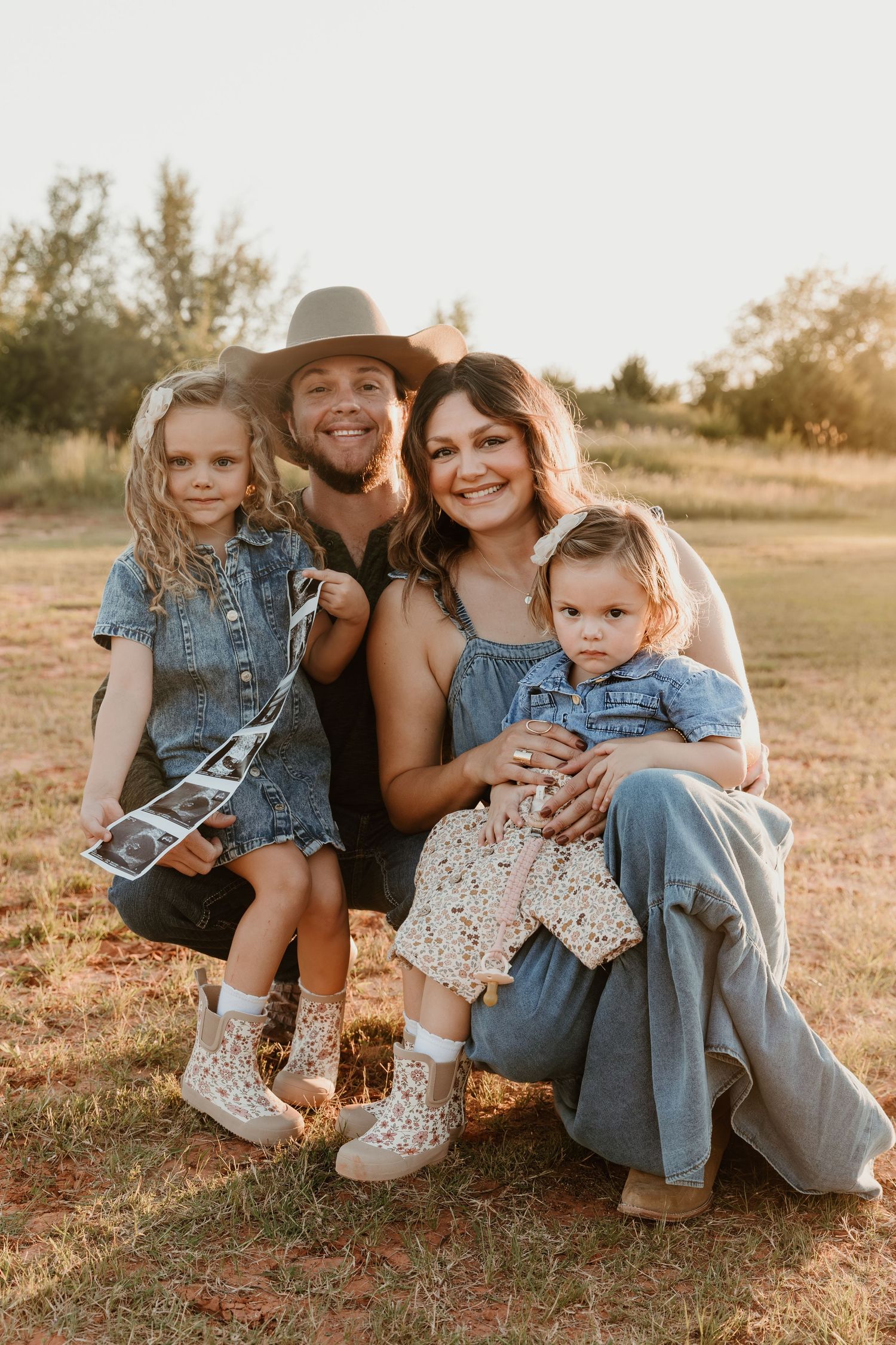 A family sits together in a grassy field at sunset wearing coordinating denim and neutral colored outfits.