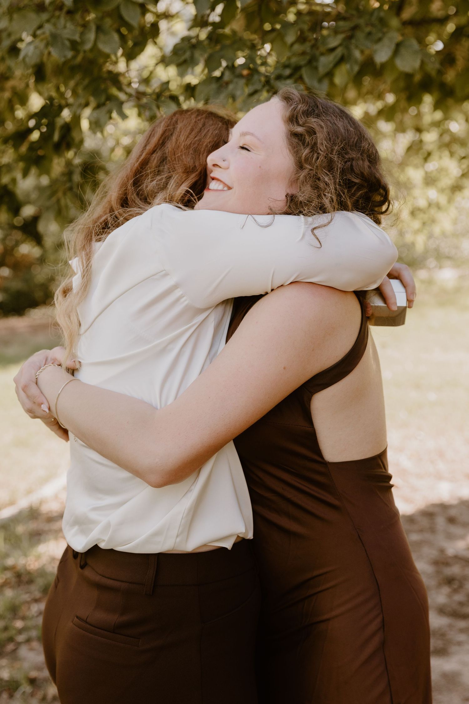 Two people share a warm embrace outdoors under dappled sunlight and tree branches.