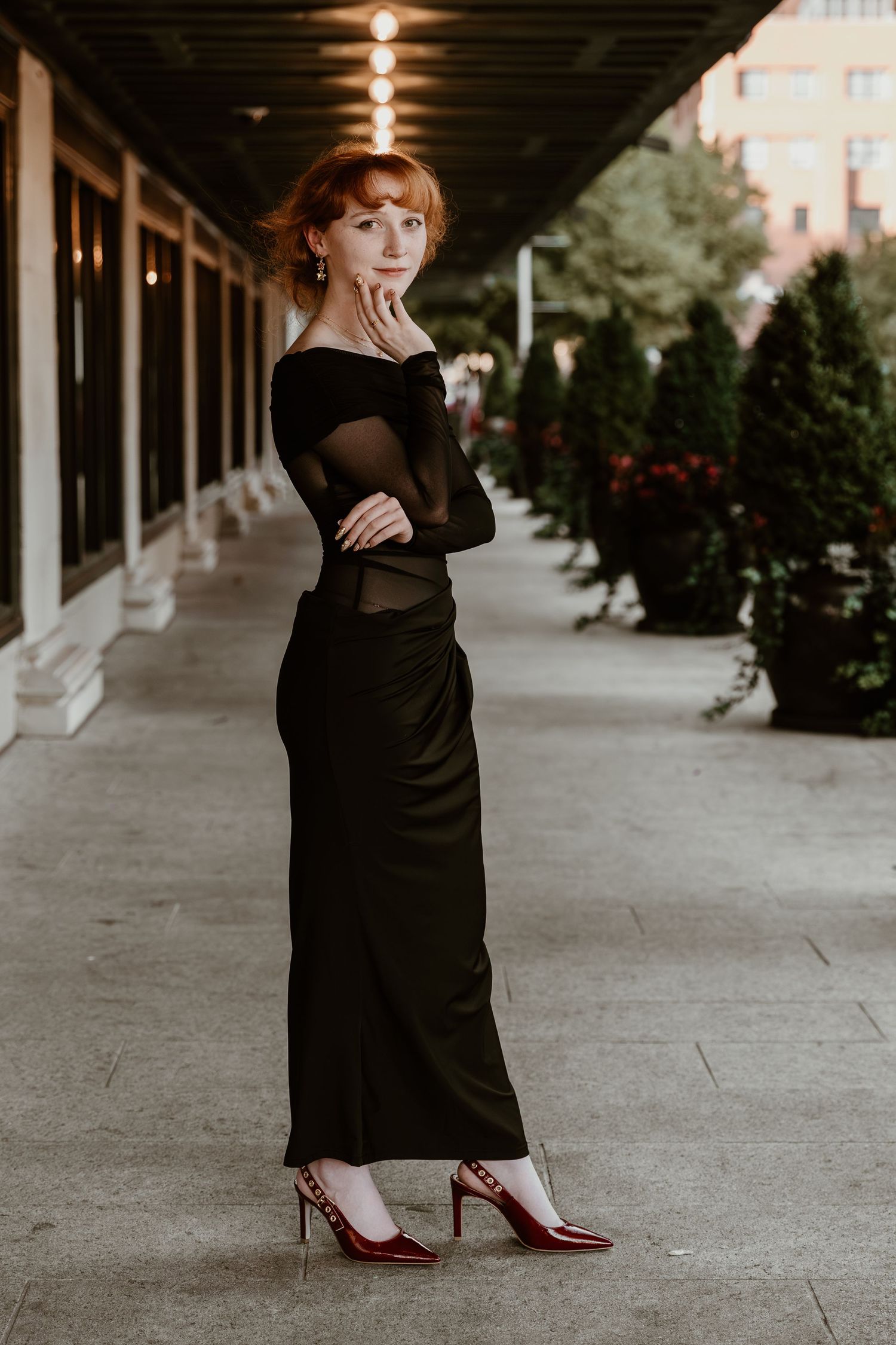 Elegant figure in a black mesh long sleeve dress and red heels poses in a covered outdoor walkway with ambient lighting.