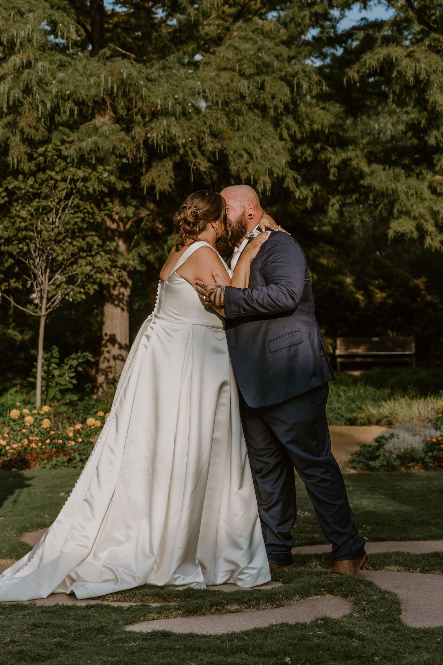 Romantic outdoor garden wedding photograph of a couple embracing in front of evergreen trees on a sunny day.