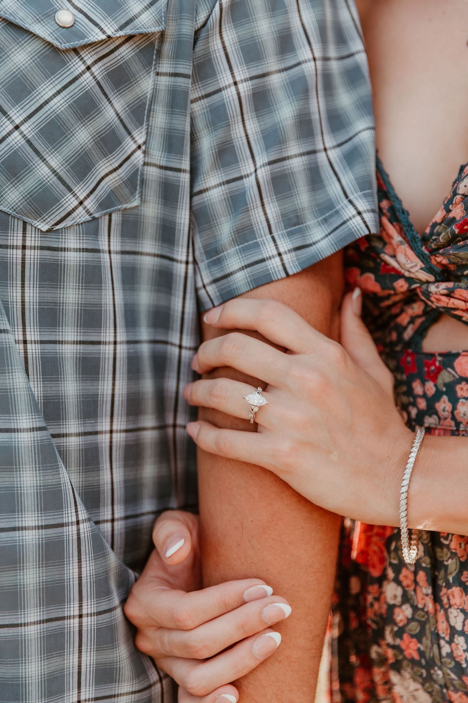 Close up of couple's hands embracing, with an engagement ring and bracelet visible against plaid shirt and floral dress.