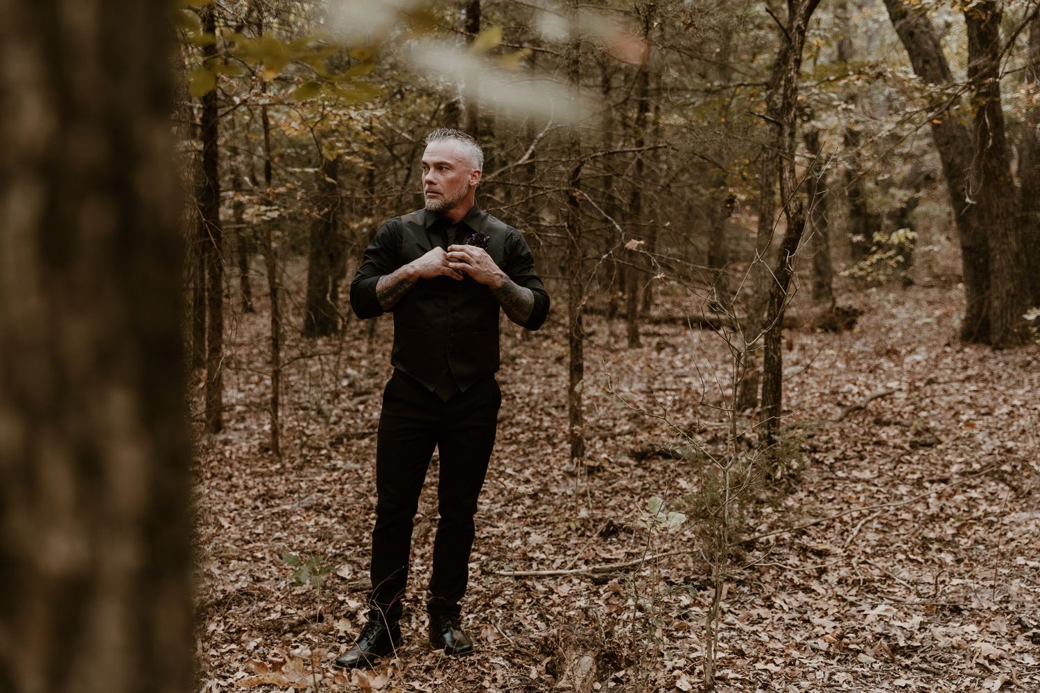A person stands in a wooded forest area during autumn with fallen leaves covering the ground.