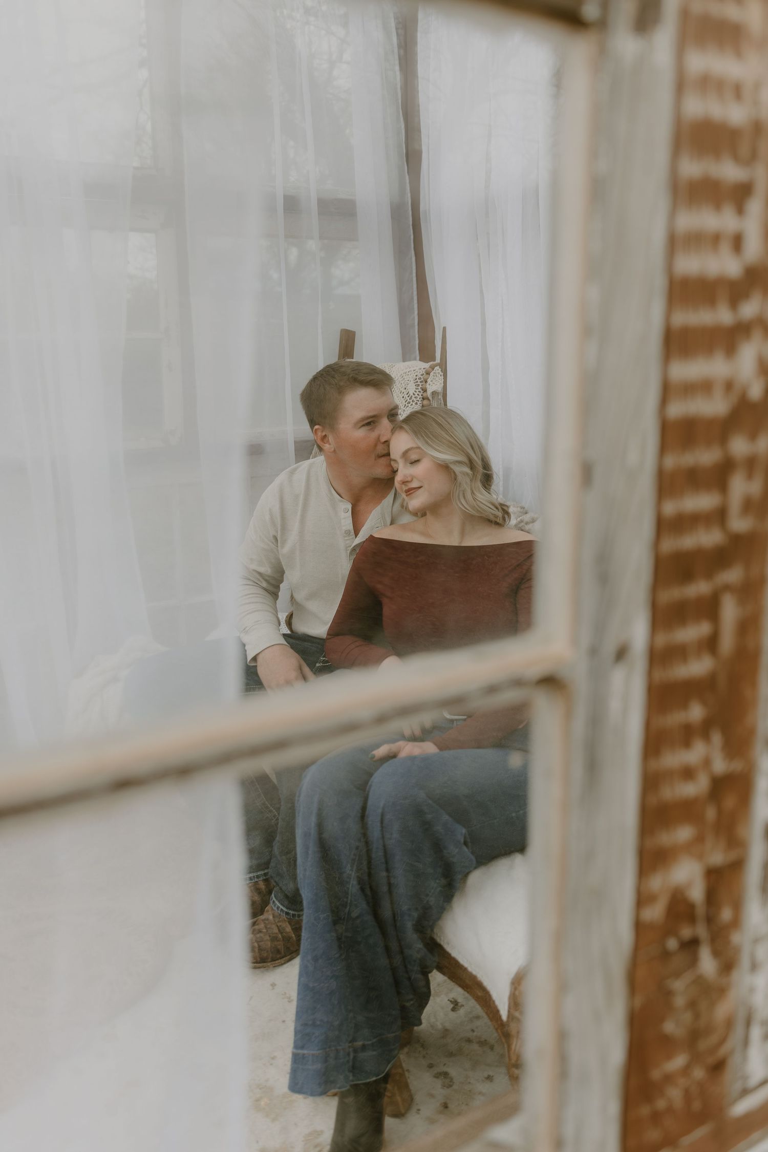 A romantic couple embraces and shares a tender moment viewed through a window with soft lighting.
