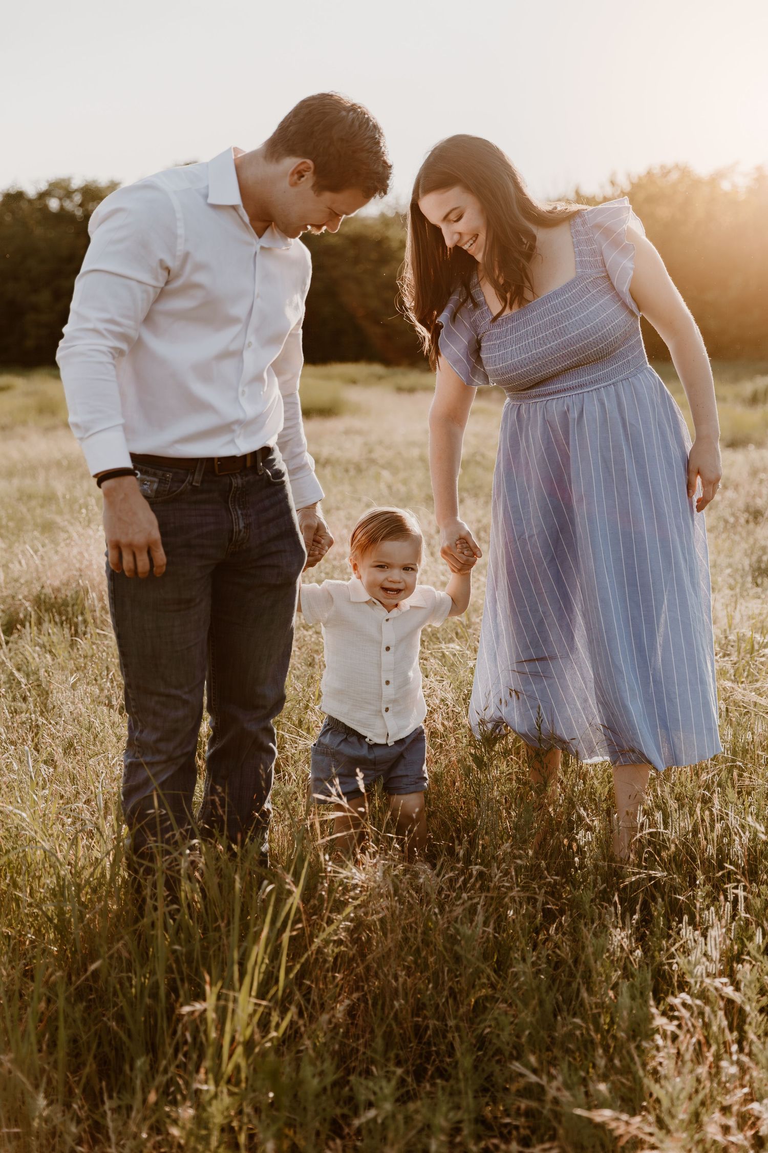 A young couple and their toddler hold hands in a sun-lit meadow during golden hour.