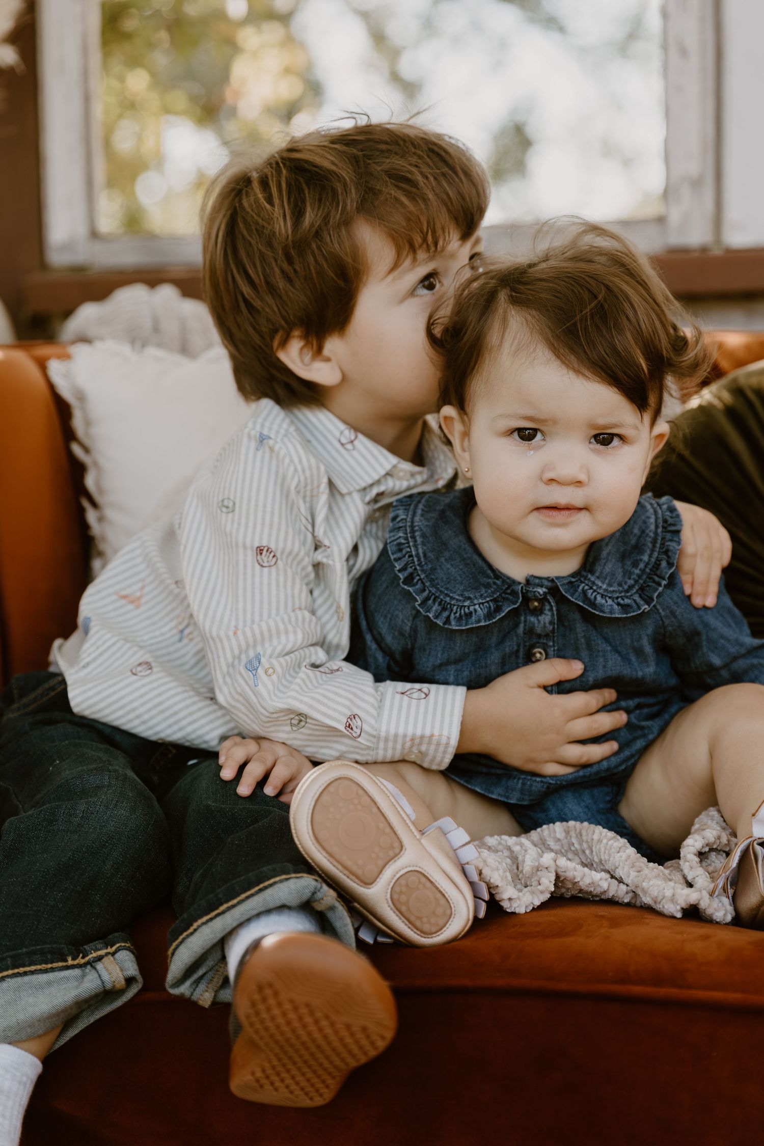 Two young children in casual attire sit together on a brown leather couch in a warm-toned indoor setting.