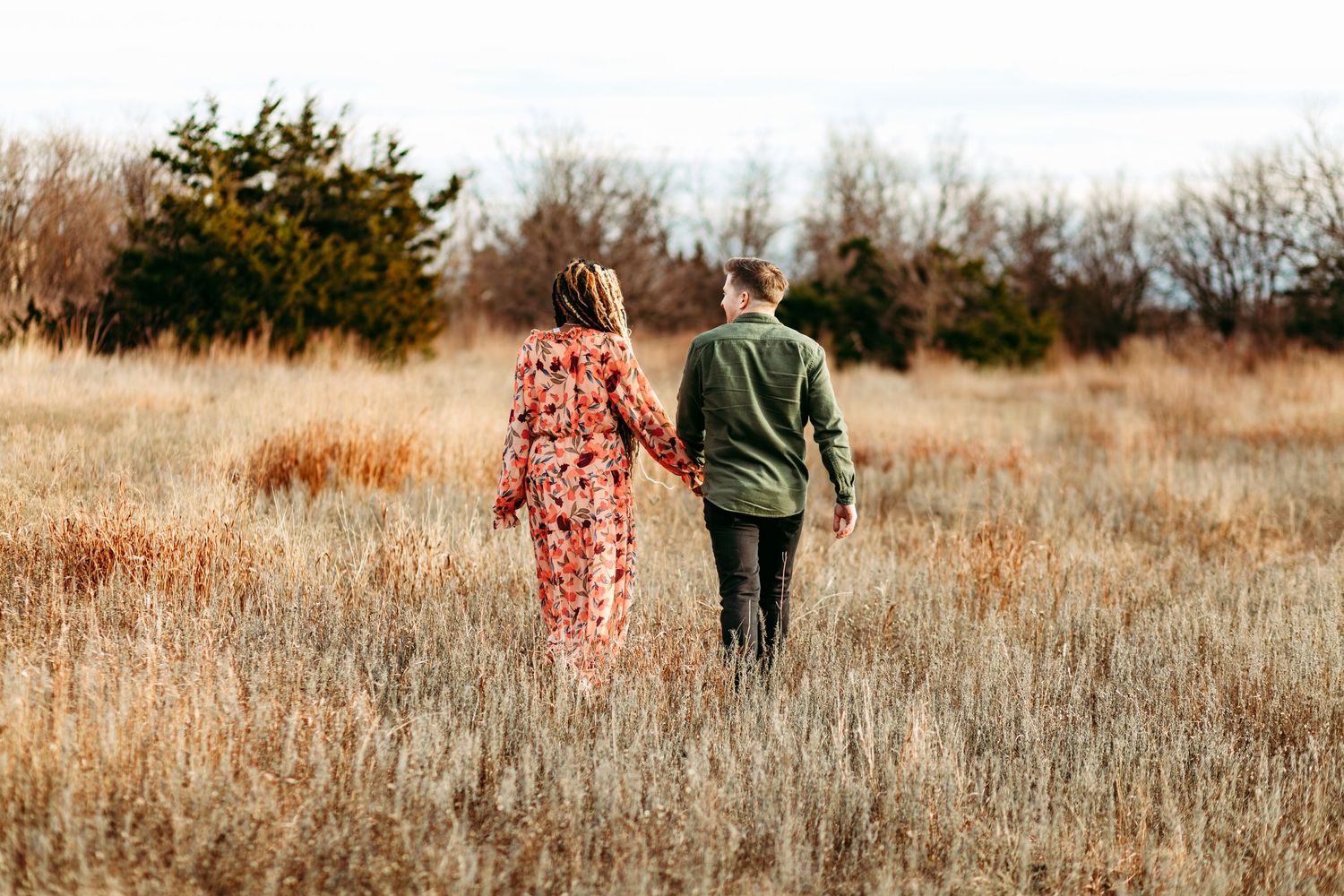 A couple walks hand in hand through tall grass in a golden field at sunset.