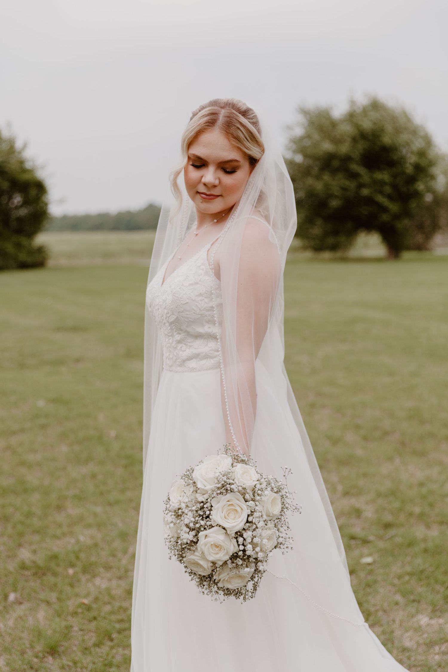 A bride in a flowing white wedding dress holds a delicate bouquet of white roses and baby's breath in a grassy field.