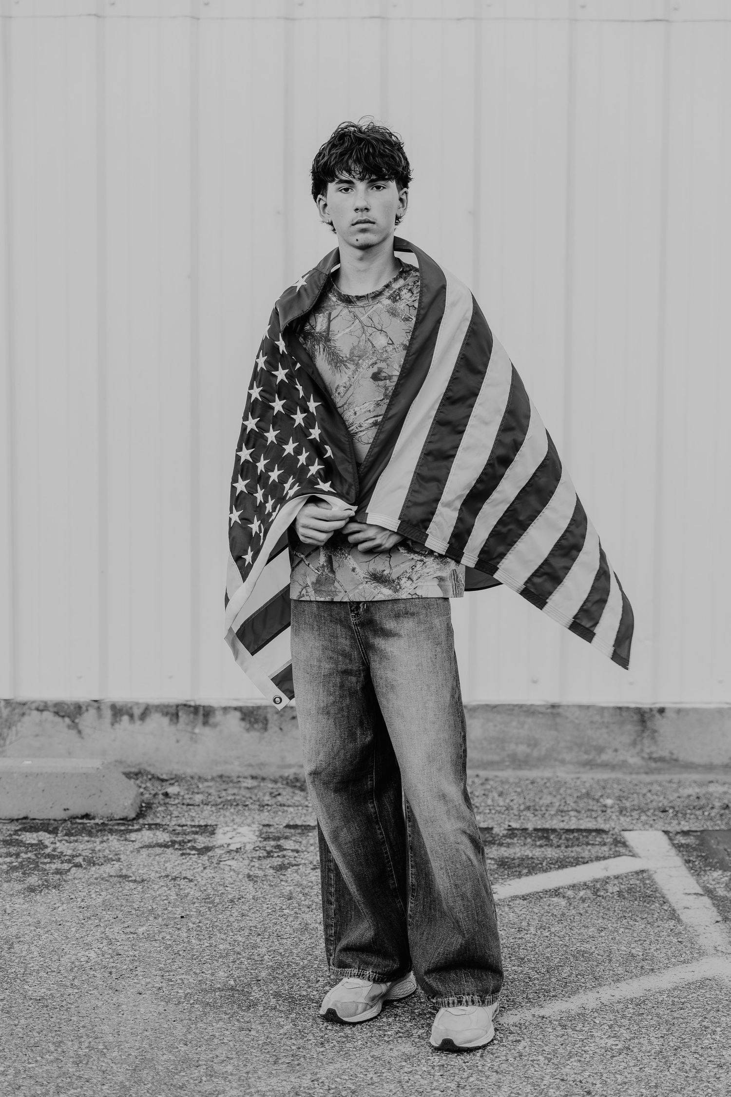A black and white photo series shows a tattooed figure draped in an American flag against a plain wall.