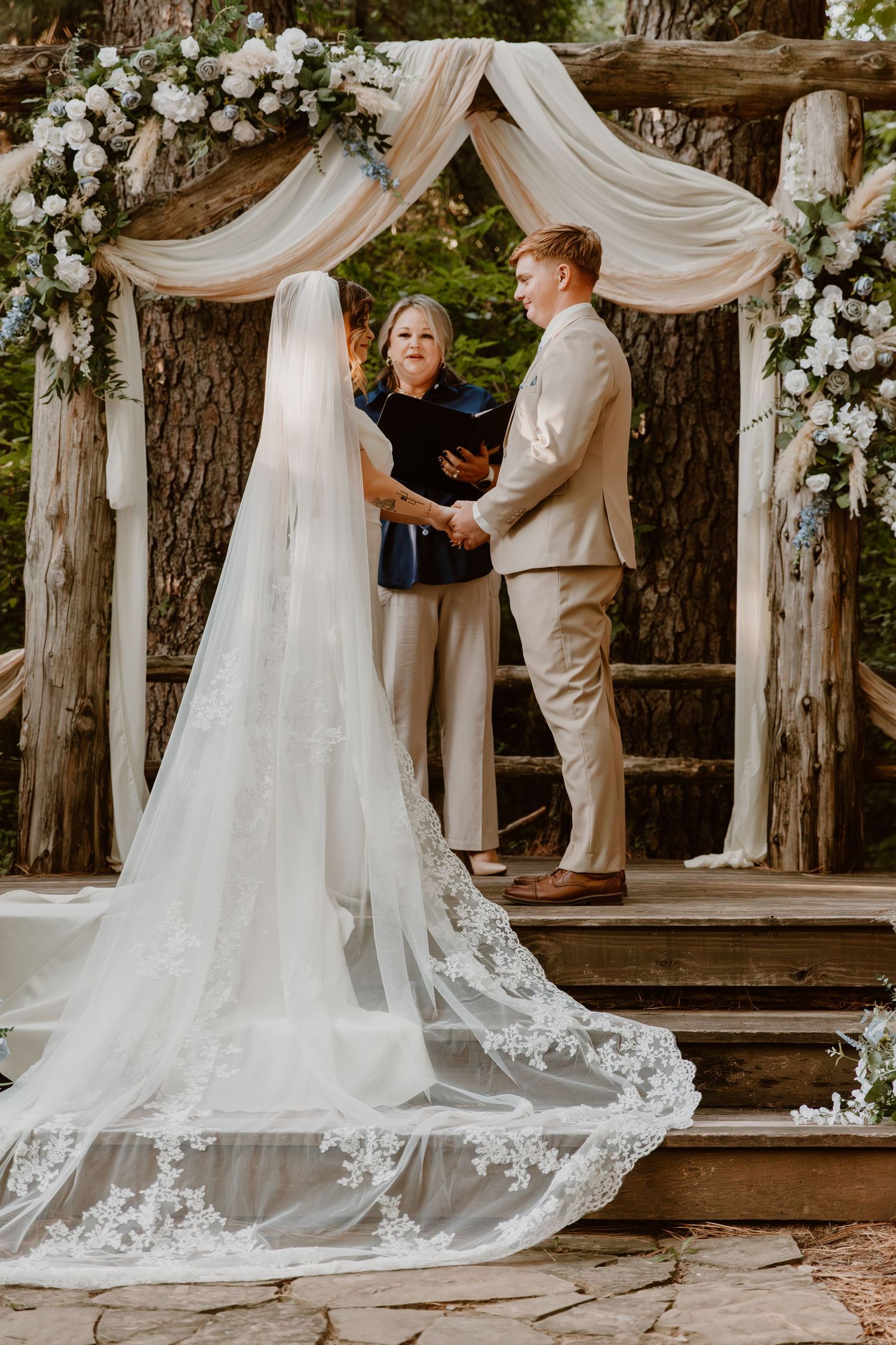 A couple exchanges vows under a draped floral wedding arch in a rustic outdoor woodland ceremony setting.
