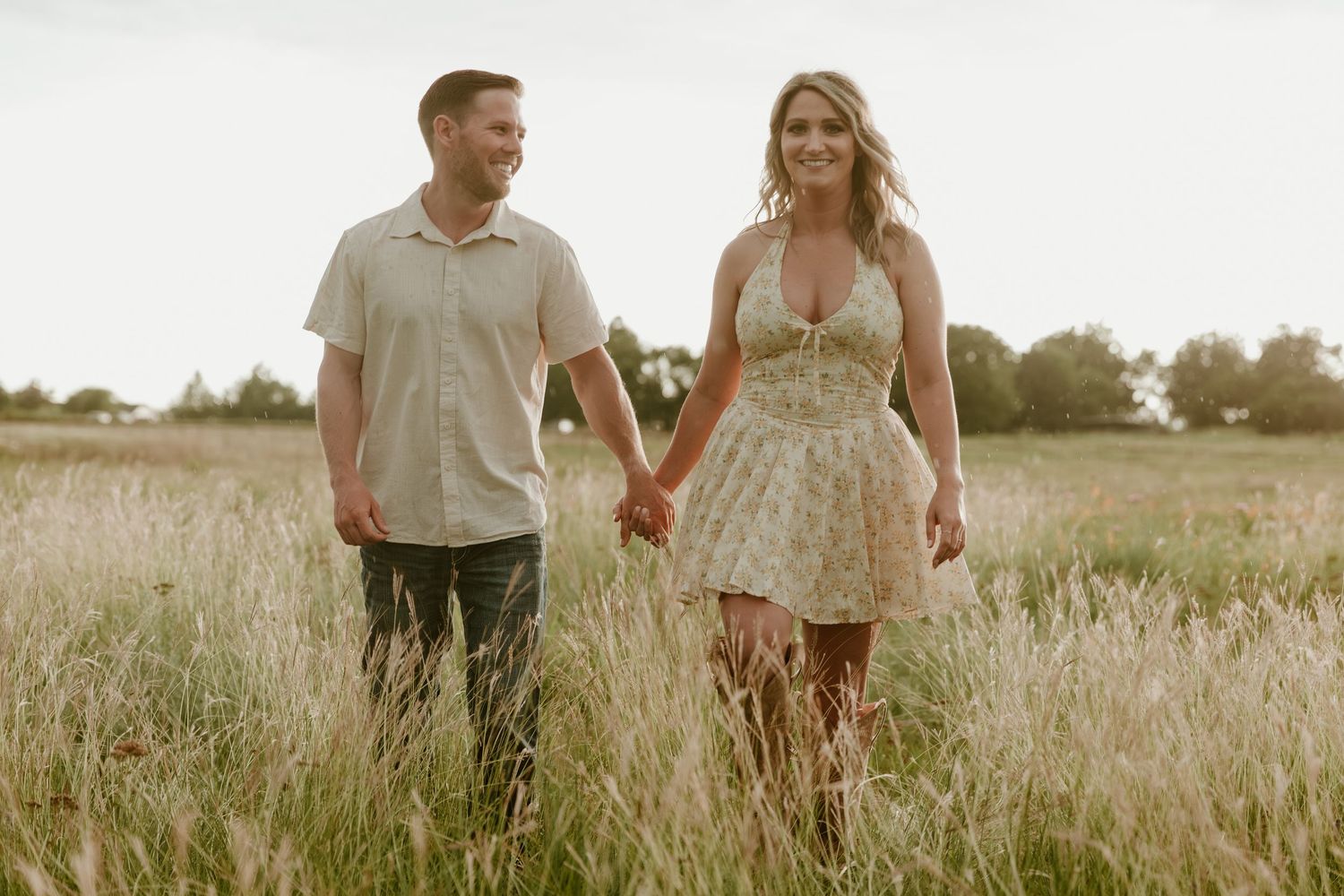 A couple walks hand in hand through a sun-drenched meadow at golden hour, creating a romantic and dreamy scene.