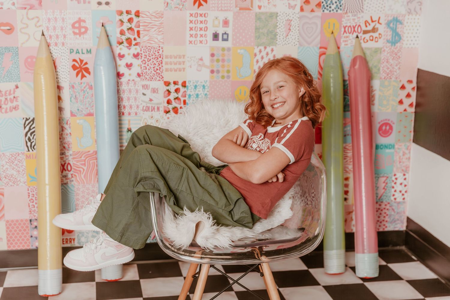 A cheerful redhead sits curled up in a retro chair against floral wallpaper with giant decorative colored pencils.