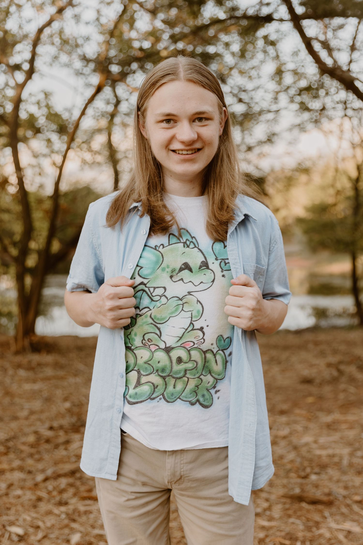 A person wearing a light blue button-up shirt and graphic tee stands outdoors among trees in warm lighting.