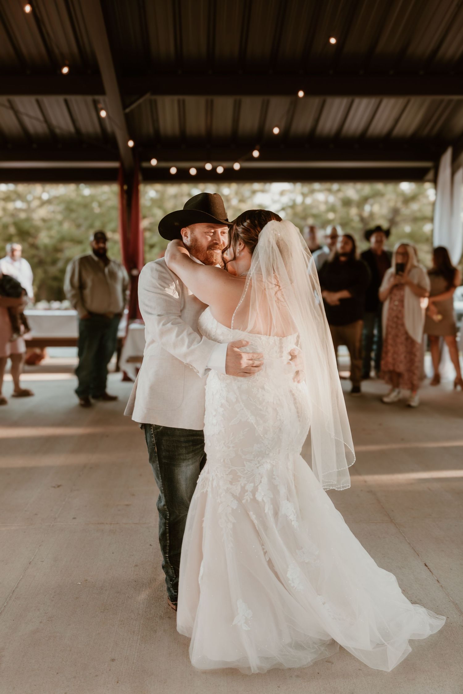 A couple shares their first dance at a rustic covered pavilion venue decorated with string lights.